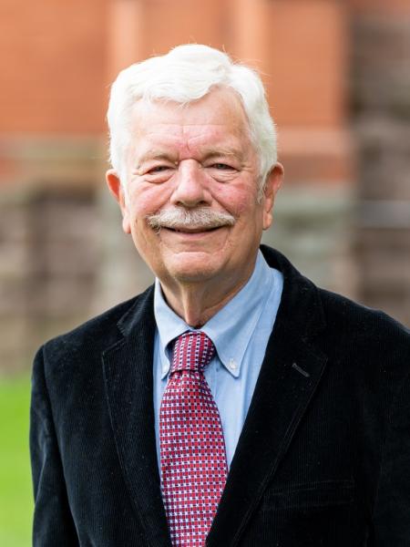 Alan Hayes in dark blue jacket and light blue shirt plus red tie in front of red brick wall at Wycliffe College